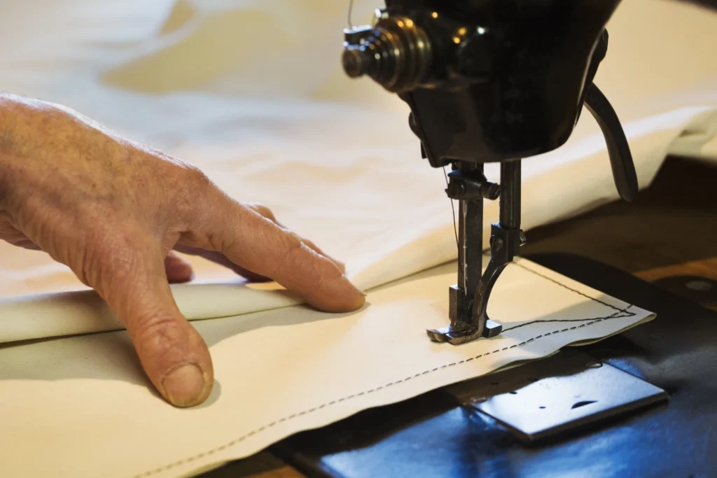 Bilde av en hånd til en seilmaker. Seilmakeren syr et hvitt seil.