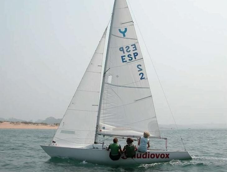 Bildet viser en seilbåtklasse med navn yngling. Denne båten har hvite seil med flere blå logoer på. Tre personer seiler langs kysten på sommeren.